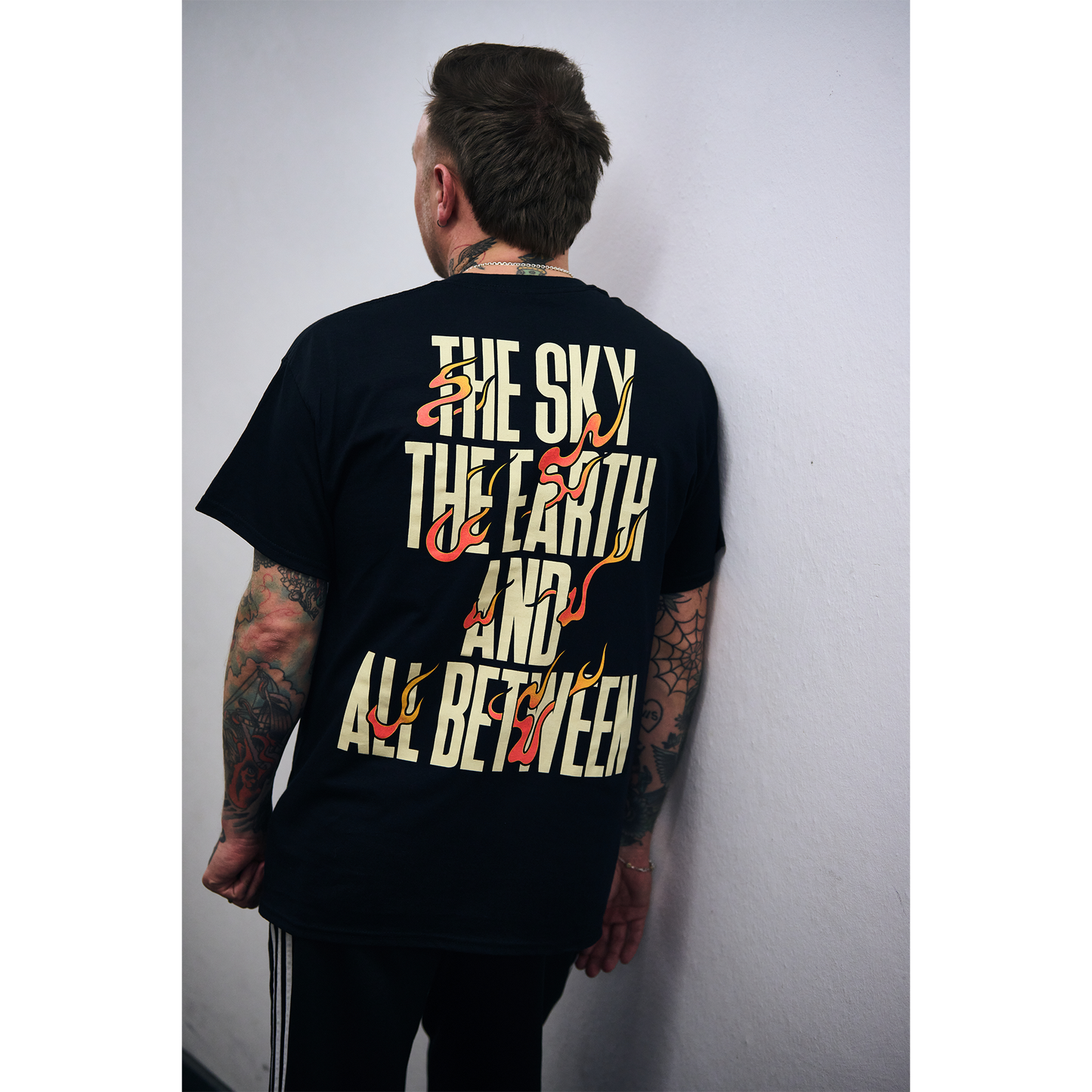 Man wearing a black short-sleeve t-shirt with fiery text design reading ‘The Sky, The Earth and All Between’ across the back