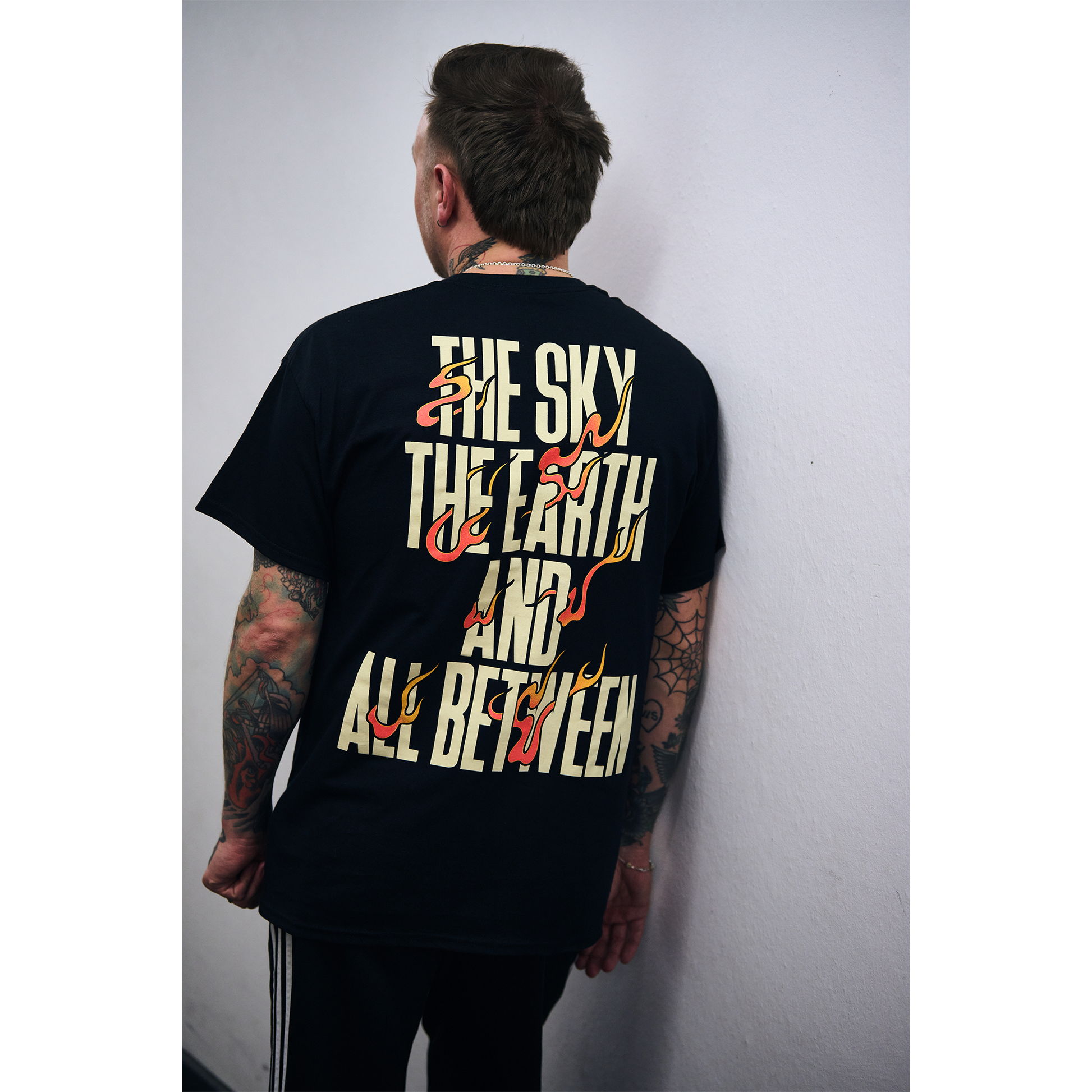 Man wearing a black short-sleeve t-shirt with fiery text design reading ‘The Sky, The Earth and All Between’ across the back