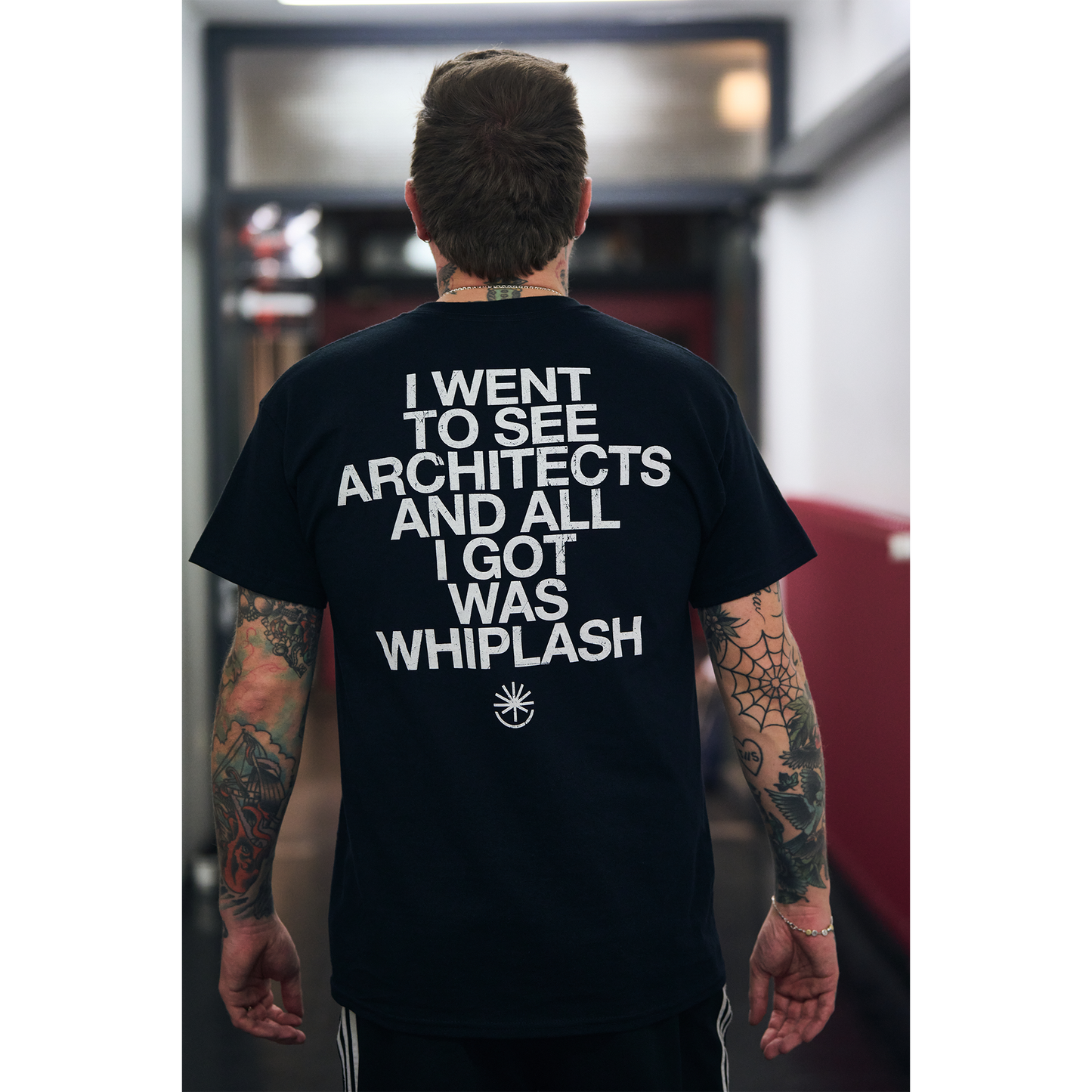 Man wearing a black unisex short-sleeve t-shirt featuring white vintage-style text that reads ‘I went to see Architects and all I got was Whiplash’ across the back