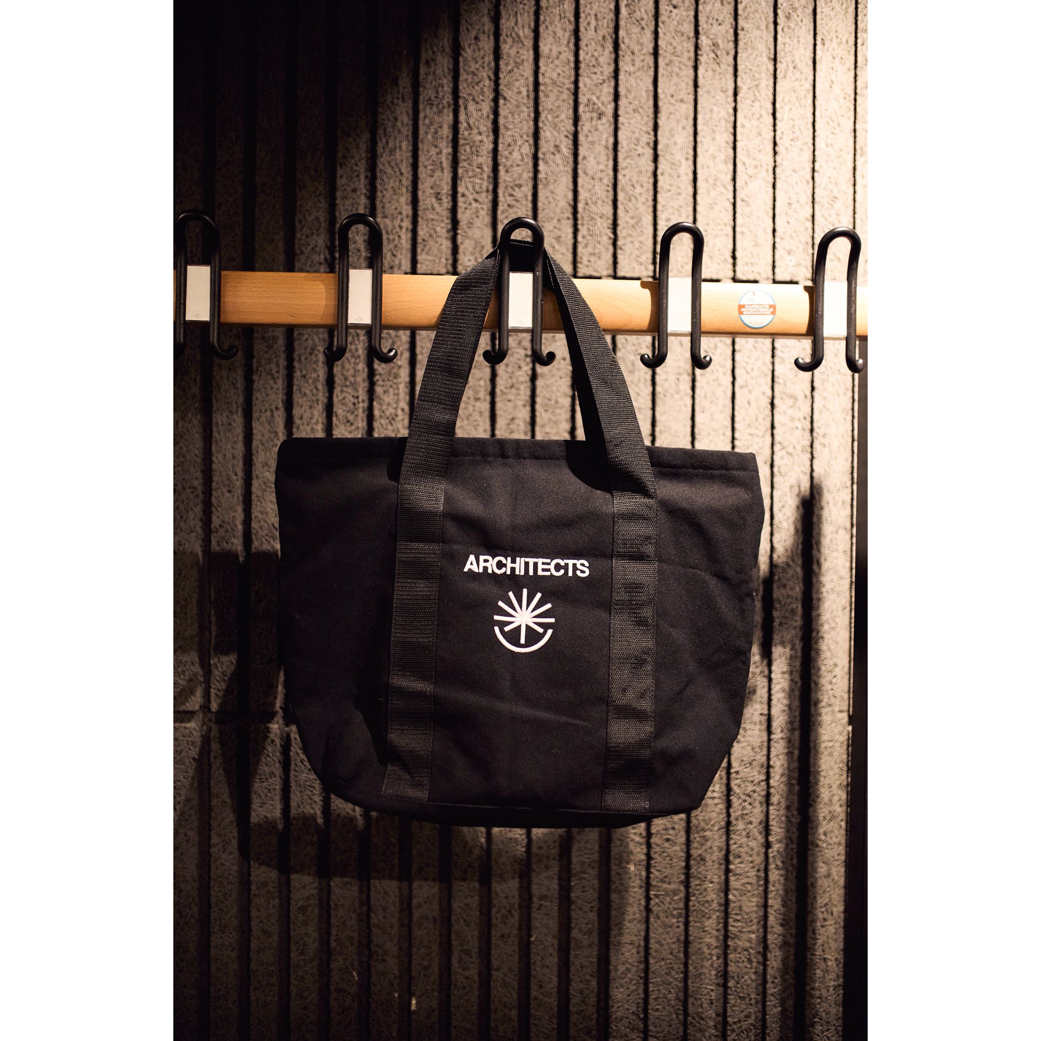 Black bag with 'ARCHITECTS' logo hanging on a wooden rack against a textured wall.